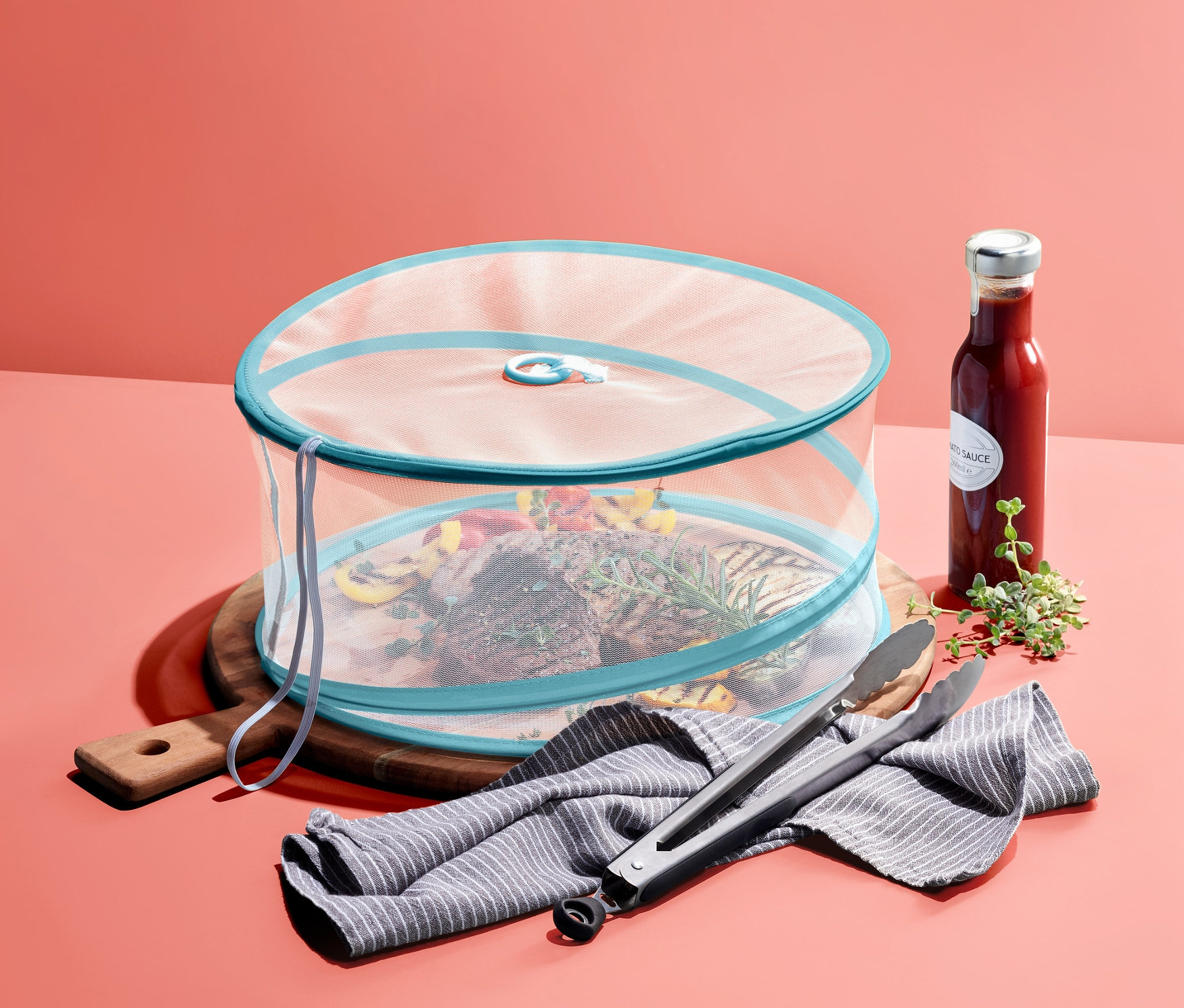 Tahta bir kesme tahtası üzerindeki yiyecekler katlanabilen sineklikle kaplıdır. Yanında bir sos şişesi, bir bulaşık bezi ve bir ızgara maşası duruyor.