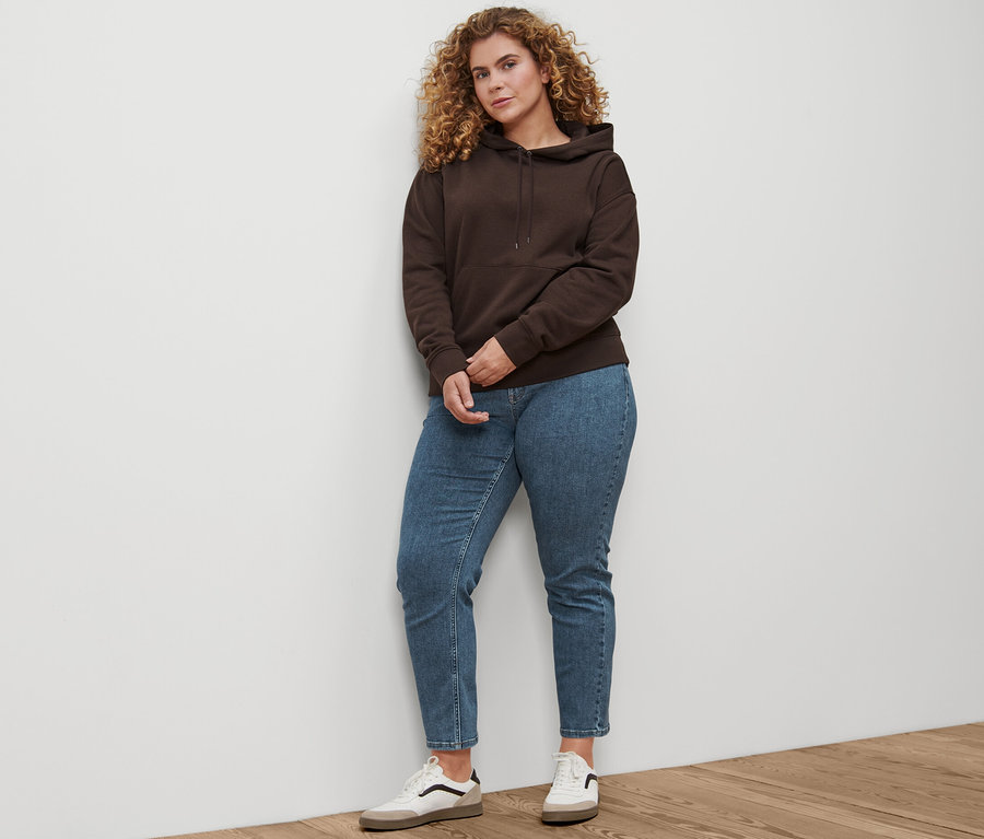 Bir kadın ayakta duruyor ve üzerinde Straight-Denim »Fit Juna«, midblue, Leder-Sneaker ve dunkelbraunen Hoodie var.