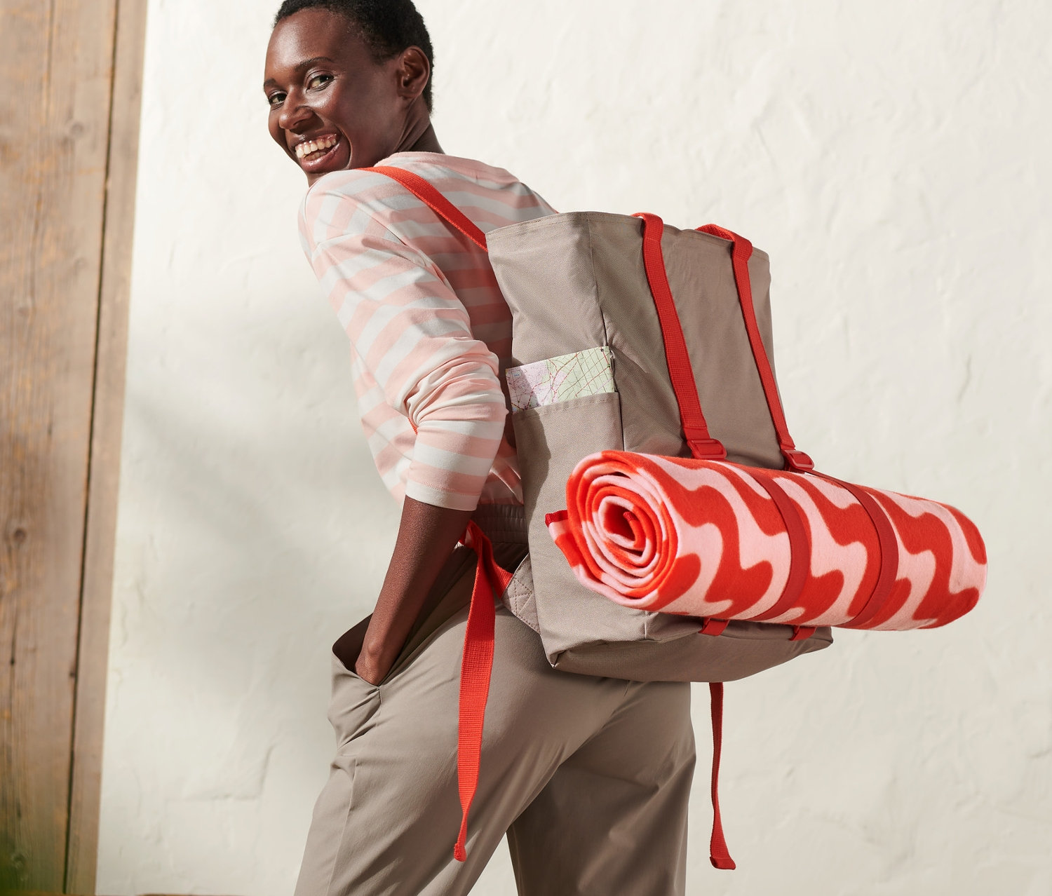 Kırmızı askılı ve kırmızı beyaz desenli rulo battaniyeli piknik sırt çantası taşıyan gülümseyen kadın.