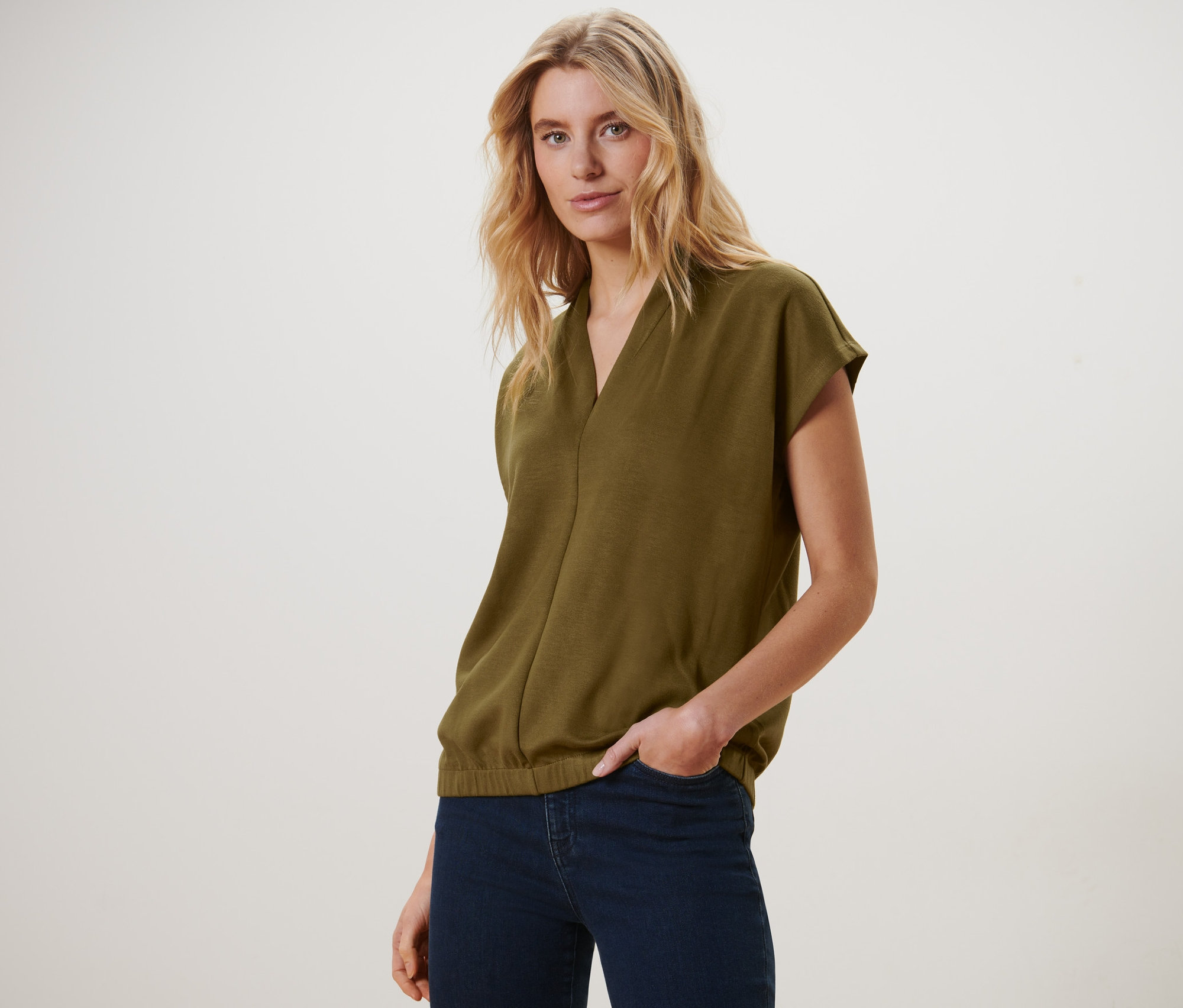 Sarışın bir kadın zeytin yeşili, V yakalı bluzu ve koyu mavi kot pantolonuyla poz veriyor.