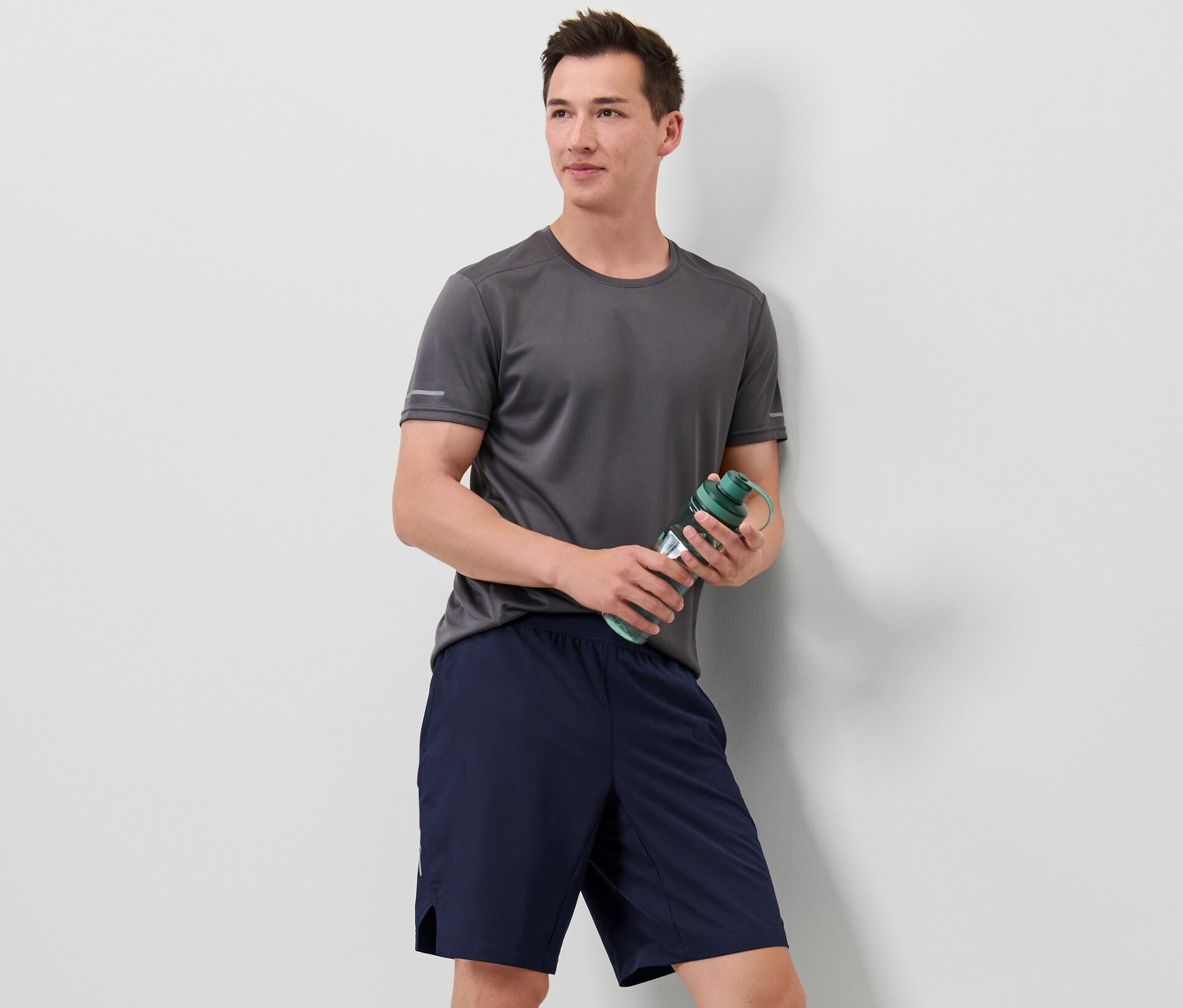 Gri tişörtlü bir adam yeşil su şişesi tutuyor ve spor şortu giyiyor.