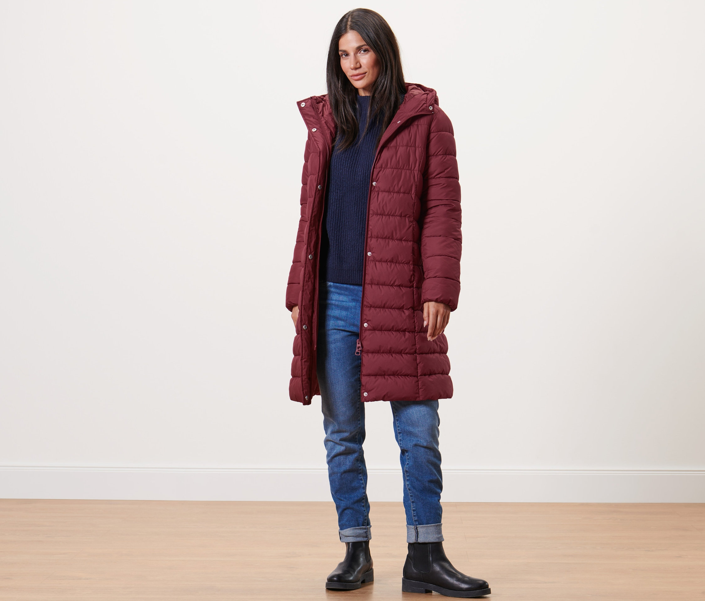 Kadın bordo kapüşonlu kapitone mont, mavi kazak, kot pantolon ve siyah bot giyiyor.