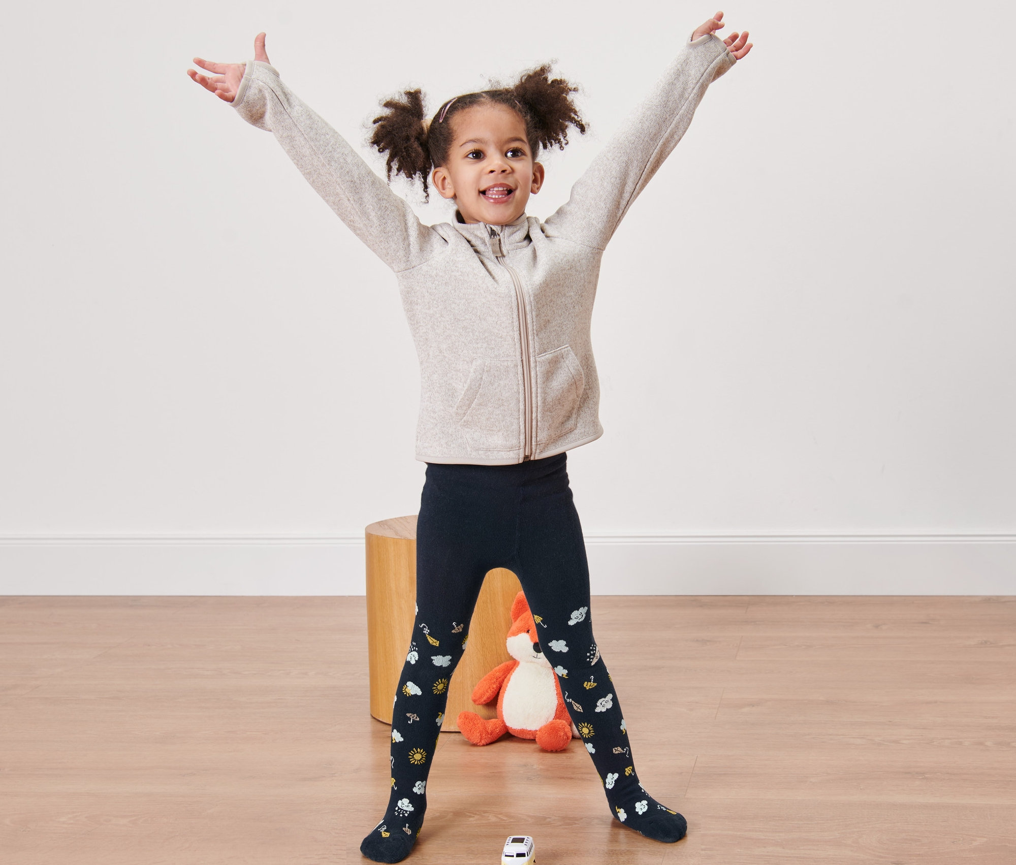 Gülümseyen bir kız çocuğu kolları açık bir şekilde ayakta duruyor ve mavi külotlu çorap giyiyor.