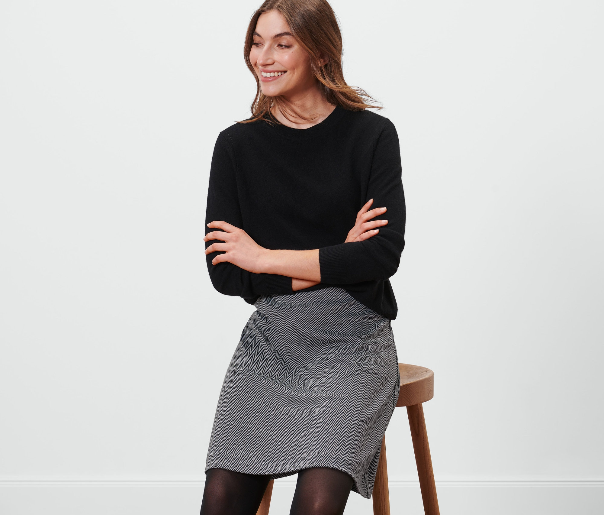 Kadın taburede oturuyor, üzerinde siyah bir kazak ve Sweat Etek var.