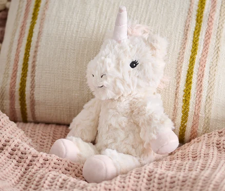 Pelüş Oyuncak »Unicorn«, bej yastığın yanında pembe örgü bir battaniyenin üzerinde oturuyor.