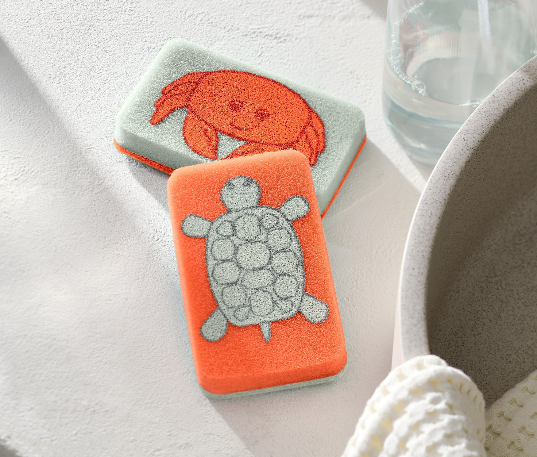 Biri yengeç, diğeri kaplumbağa desenli iki adet temizlik süngeri yan yana duruyor.