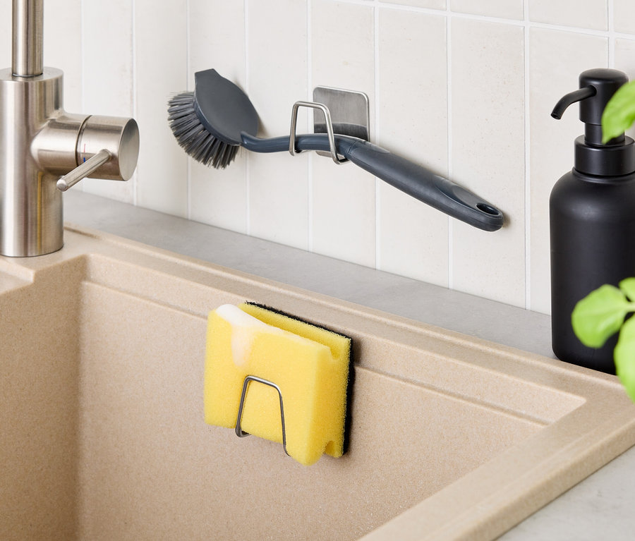 Mutfakta bej bir lavabo ve musluk var. Duvarda fırçalı bir tutucu ve lavaboda süngerli bir tutucu var. Lavabonun yanında siyah bir şişe duruyor.