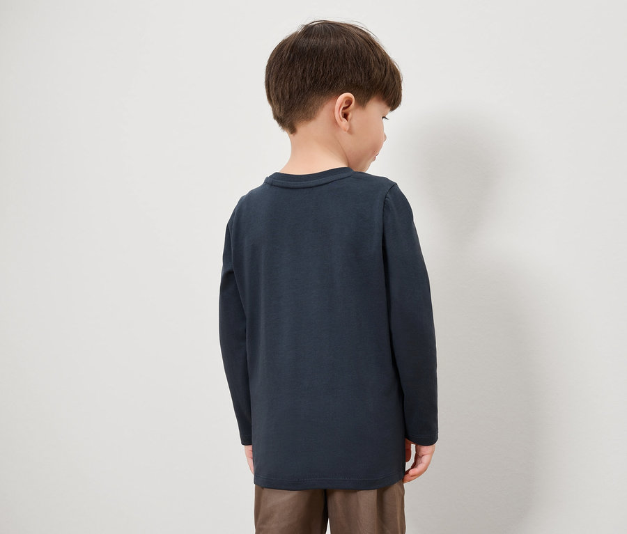 Sırtı kameraya dönük bir erkek çocuk, lacivert uzun kollu bir tişört ve kahverengi pantolon giyiyor.