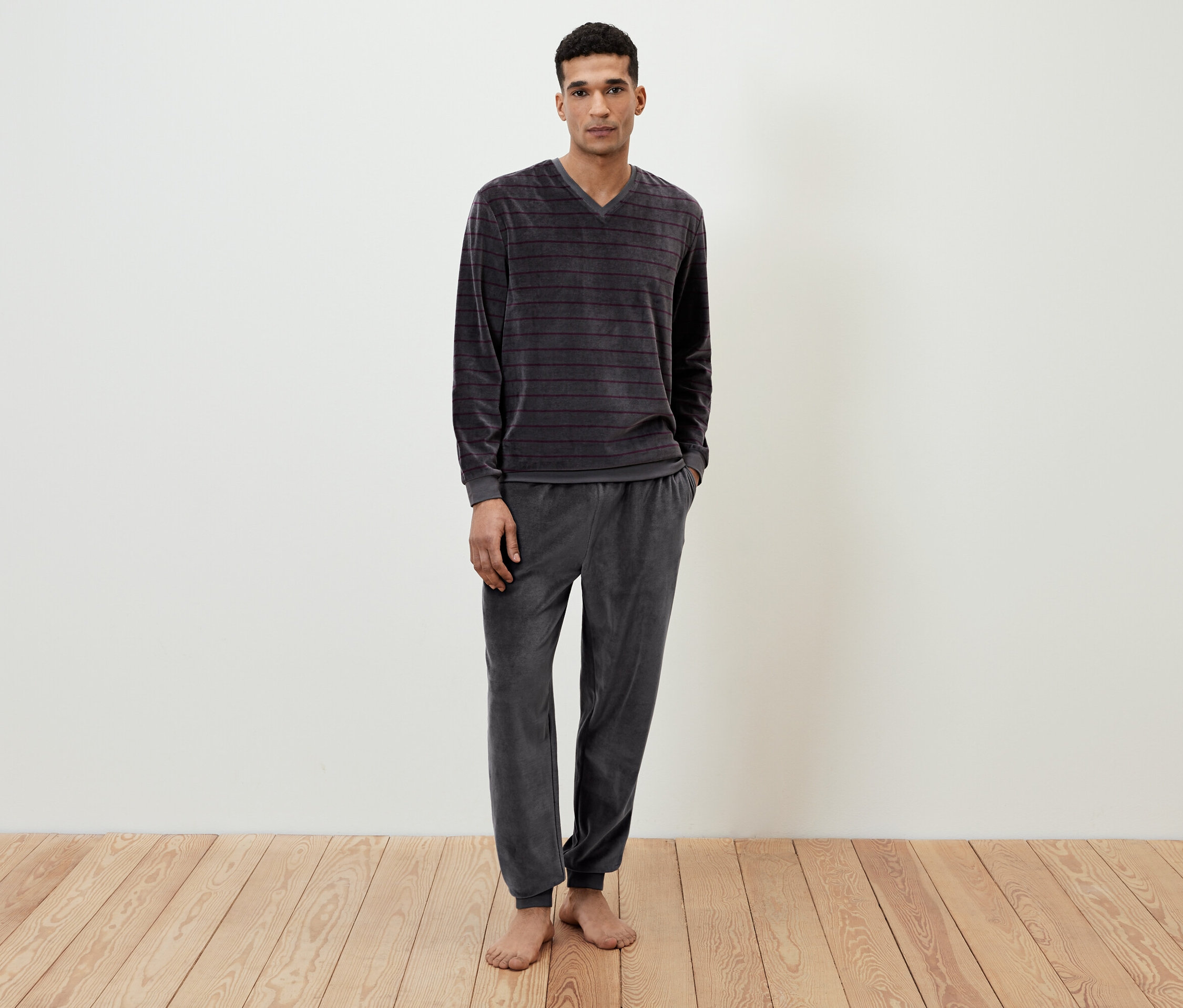Adam yerde çıplak ayakla duruyor ve gri, çizgili, kadife bir pijama giyiyor.