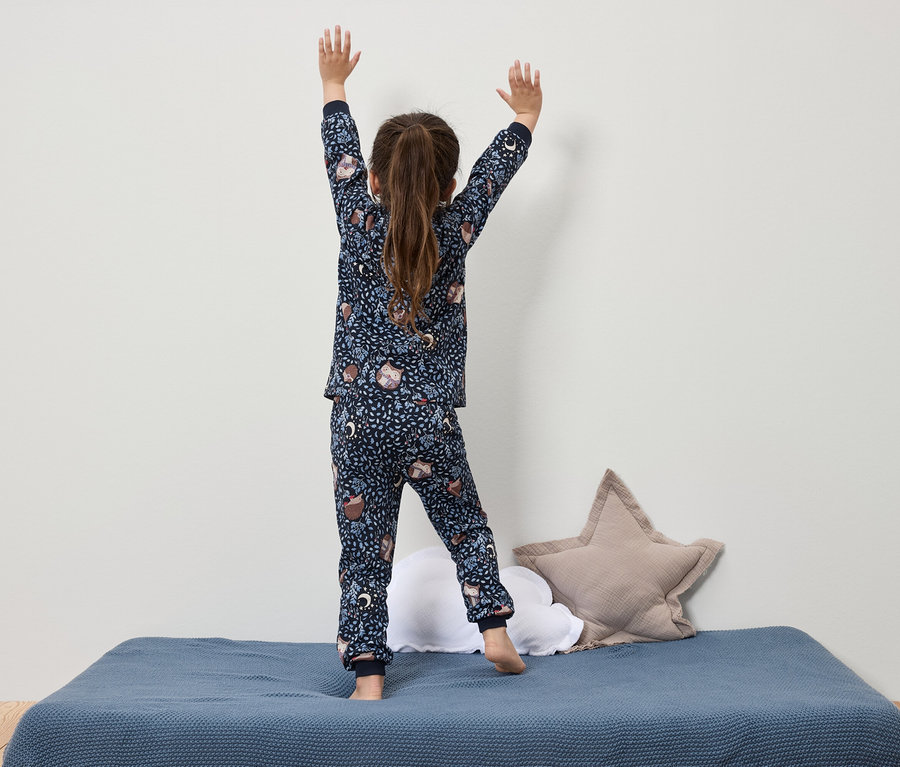 Desenli lacivert pijamalı kız, mavi bir yatakta elleri havada duruyor.