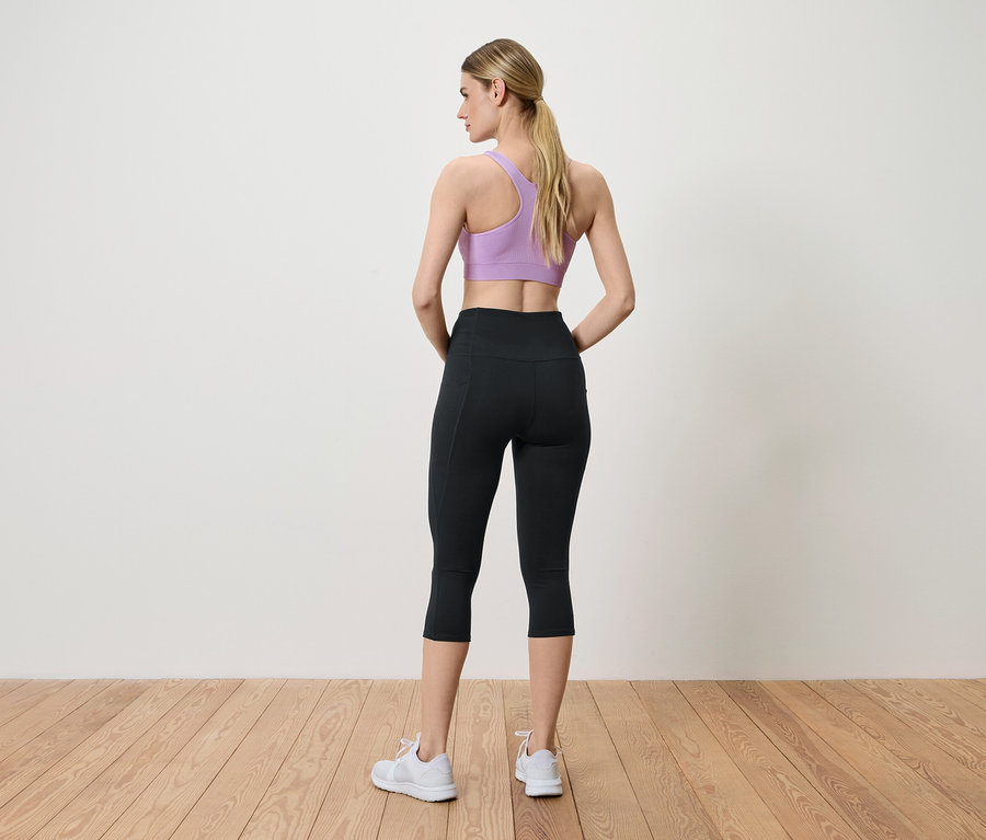 Kadın kameraya arkası dönük poz veriyor, mor spor sütyeni, siyah taytı ve beyaz spor ayakkabıları giyiyor.