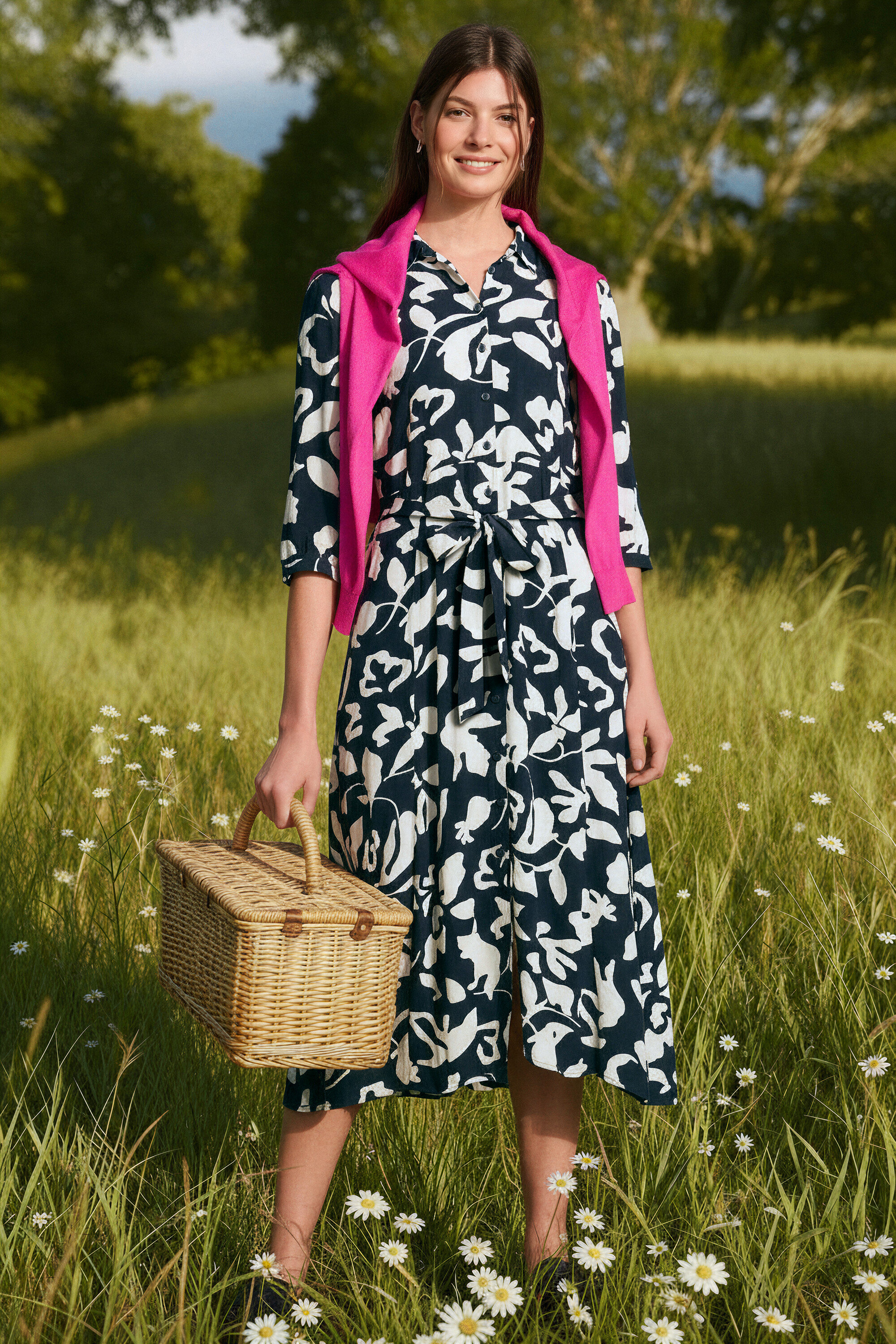 Çayırda duran ve piknik sepeti tutan bir kadın. Dalga Halka Küpe ve Feinstrick-Cardigan giyiyor.