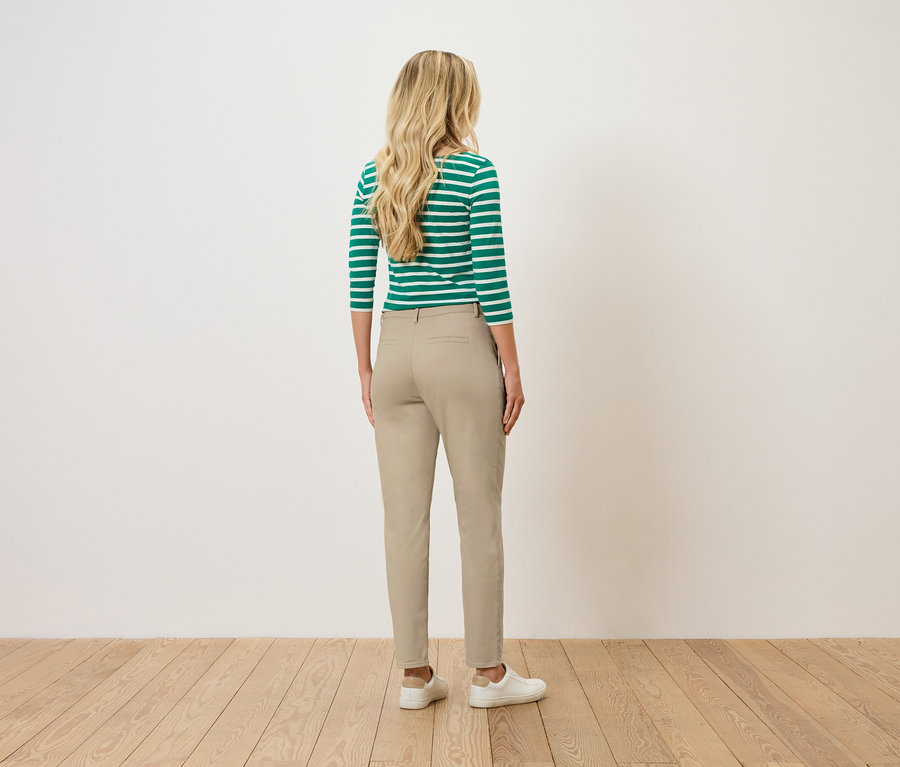 Sarışın, dalgalı saçlı bir kadın, sırtı izleyiciye dönük, yeşil ve beyaz çizgili bir üst, bej pantolon ve beyaz spor ayakkabılar giyiyor.