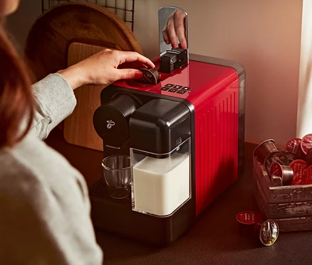 Kadın kırmızı Cafissimo milk kahve makinesine kapsül yerleştiriyor. Yanında bir bardak süt ve kapsüllerle dolu ahşap bir kutu var.
