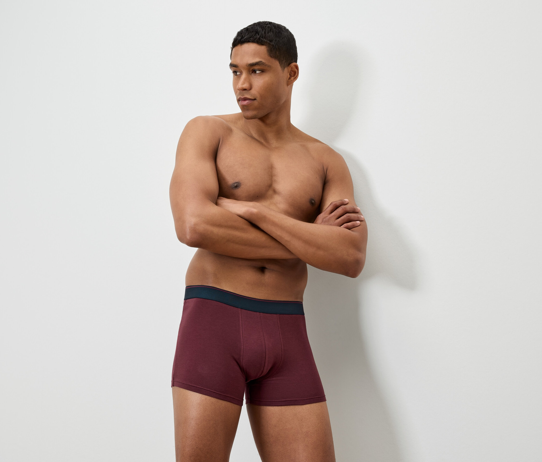 Kollarını kavuşturmuş bir adam bordo renkli boxer şort giyiyor.