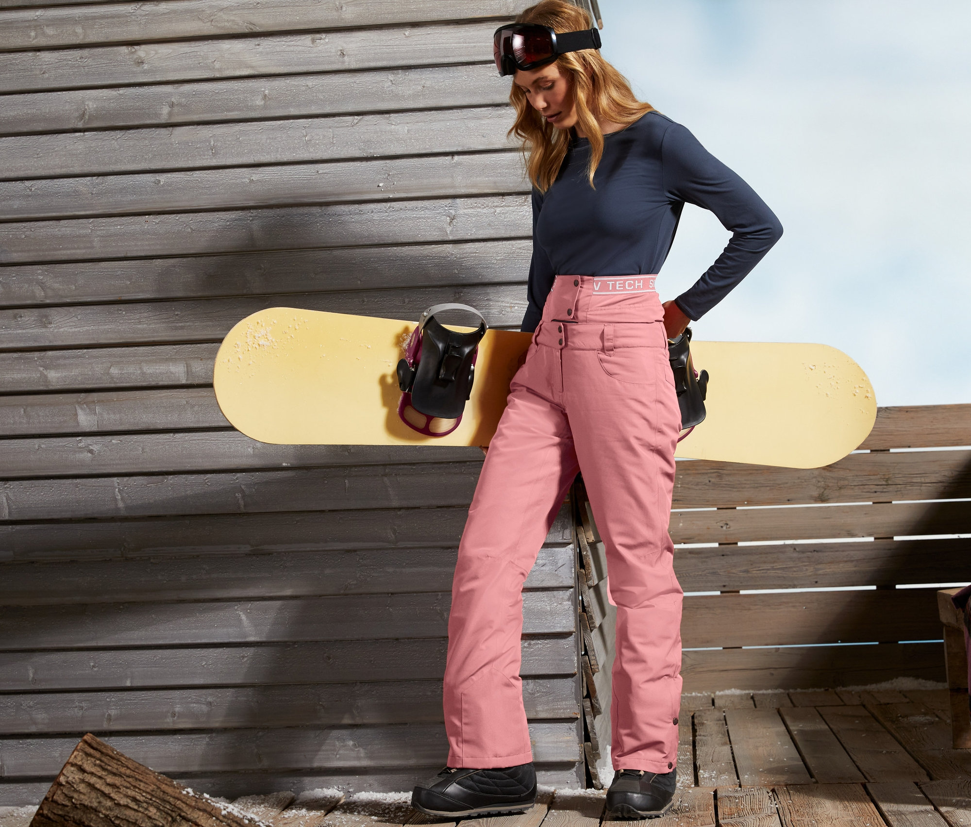 Kadın ahşap bir duvarın yanında duruyor ve bir snowboard'a yaslanıyor. Kayak ve Snowboard Pantolonu giyiyor.