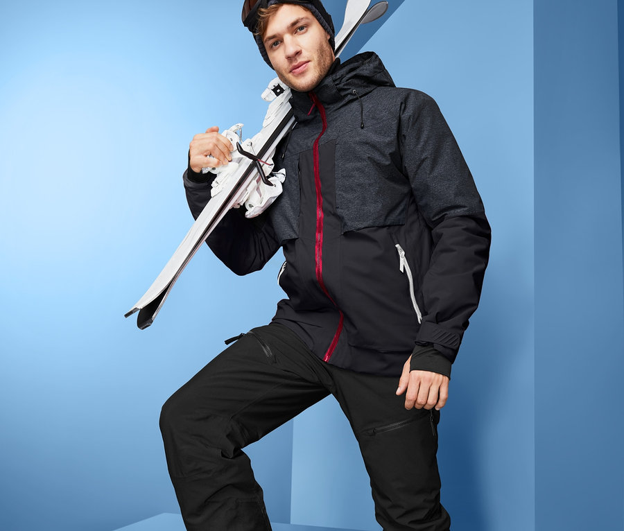 High-End Kayak Montu, kayak pantolonu, kayak ayakkabısı ve gözlüğü giyen bir adam omzunda kayak taşıyor.