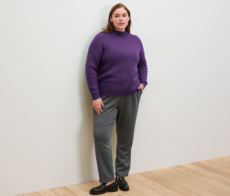Bir kadın, elleri jogger pantolonunun cebinde, üzerinde mor Yarım Boğazlı Kalın Örgülü Kazak ile ayakta duruyor.
