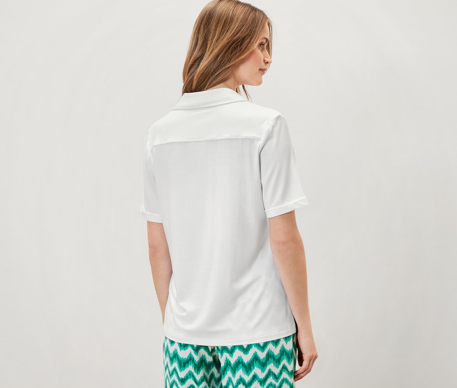 Kadın kameraya arkası dönük duruyor, beyaz düğmeli bluz ve yeşil desenli pantolon giyiyor.