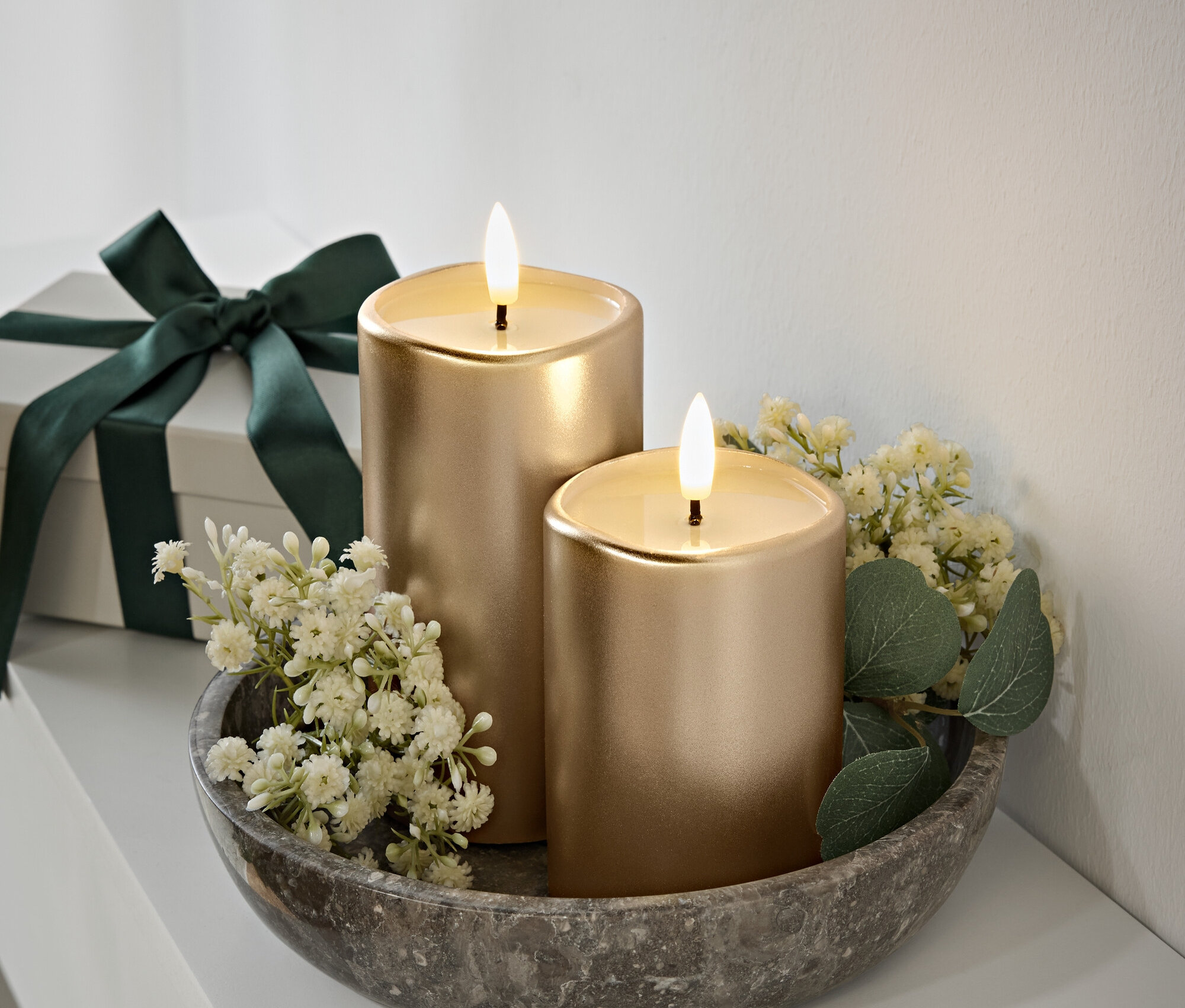 Gri taş bir kasede alevli iki altın mum, beyaz çiçekler ve yeşil yapraklar. Arka planda koyu yeşil kurdeleli hediye kutusu.