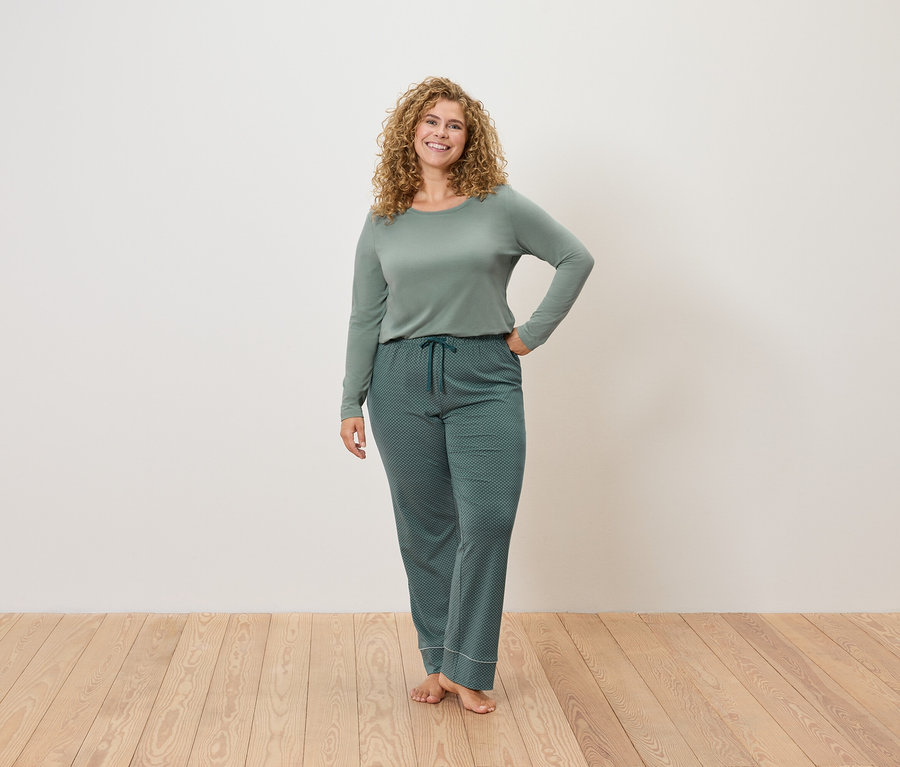 Kadın beyaz bir duvarın önünde ahşap bir zeminde yeşil pijamalarla ayakta duruyor ve poz veriyor.