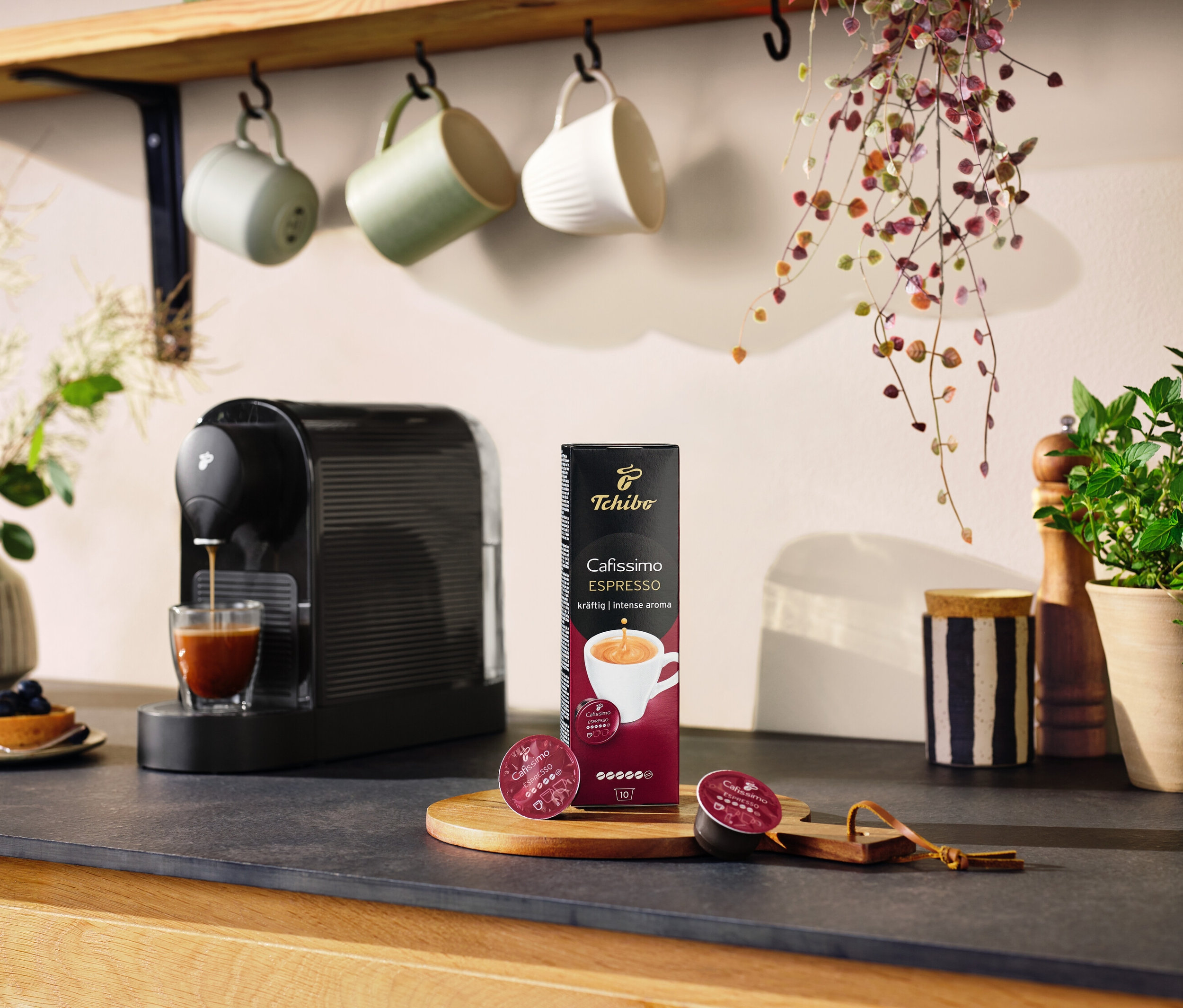 Siyah Cafissimo pure+ bir bardağa kahve yapıyor. Masanın üzerinde bir paket kapsül ve iki kapsül duruyor.