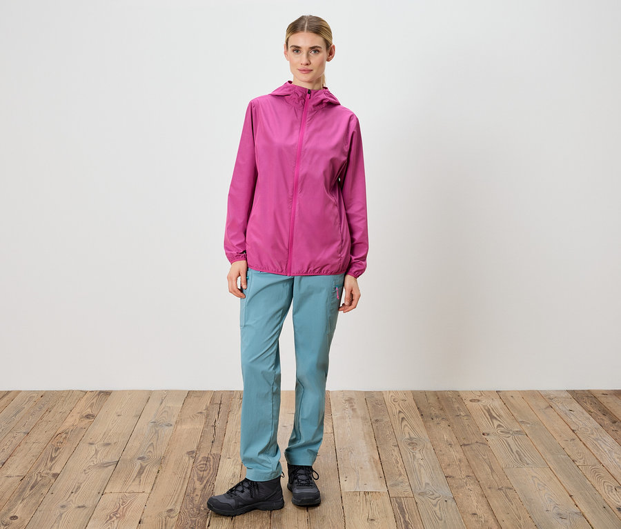 Kadın beyaz bir duvarın önünde duruyor, pembe bir ceket, mavi pantolon ve siyah ayakkabılar giyiyor.