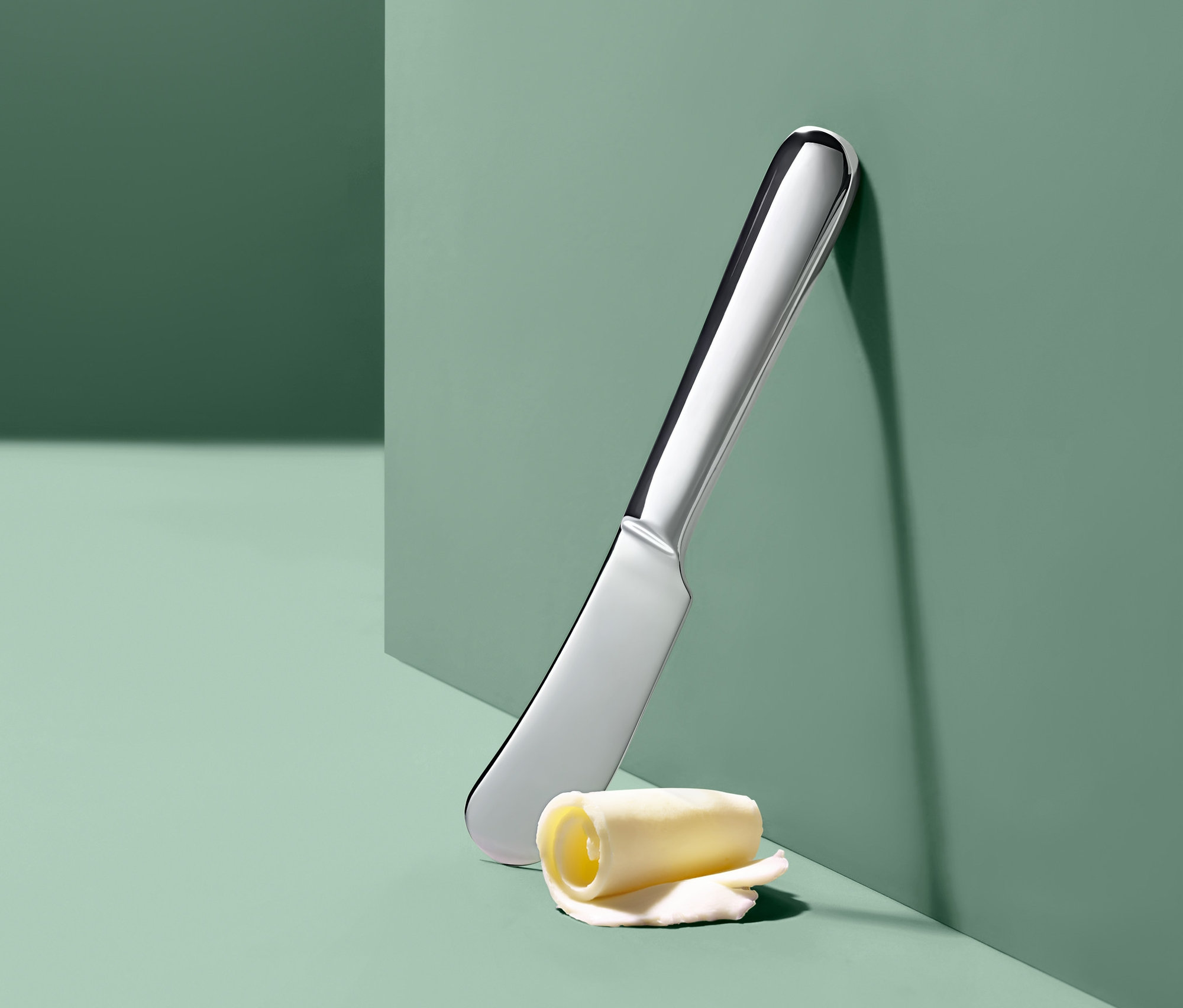 Gümüş renkli bir tereyağı bıçağı ve bir tereyağı rulosu yeşil bir duvara yaslanmış.