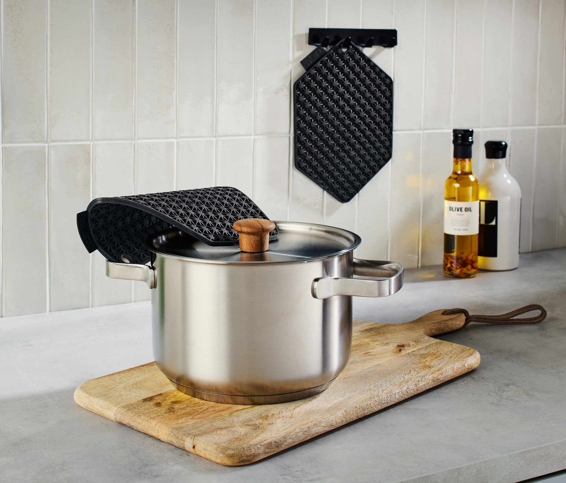 Ahşap kesme tahtası üzerinde kapaklı paslanmaz çelik tencere, yanında mutfak gereçleri olan yeşil bir sürahi ve siyah 3'ü 1 arada mutfak yardımcısı.