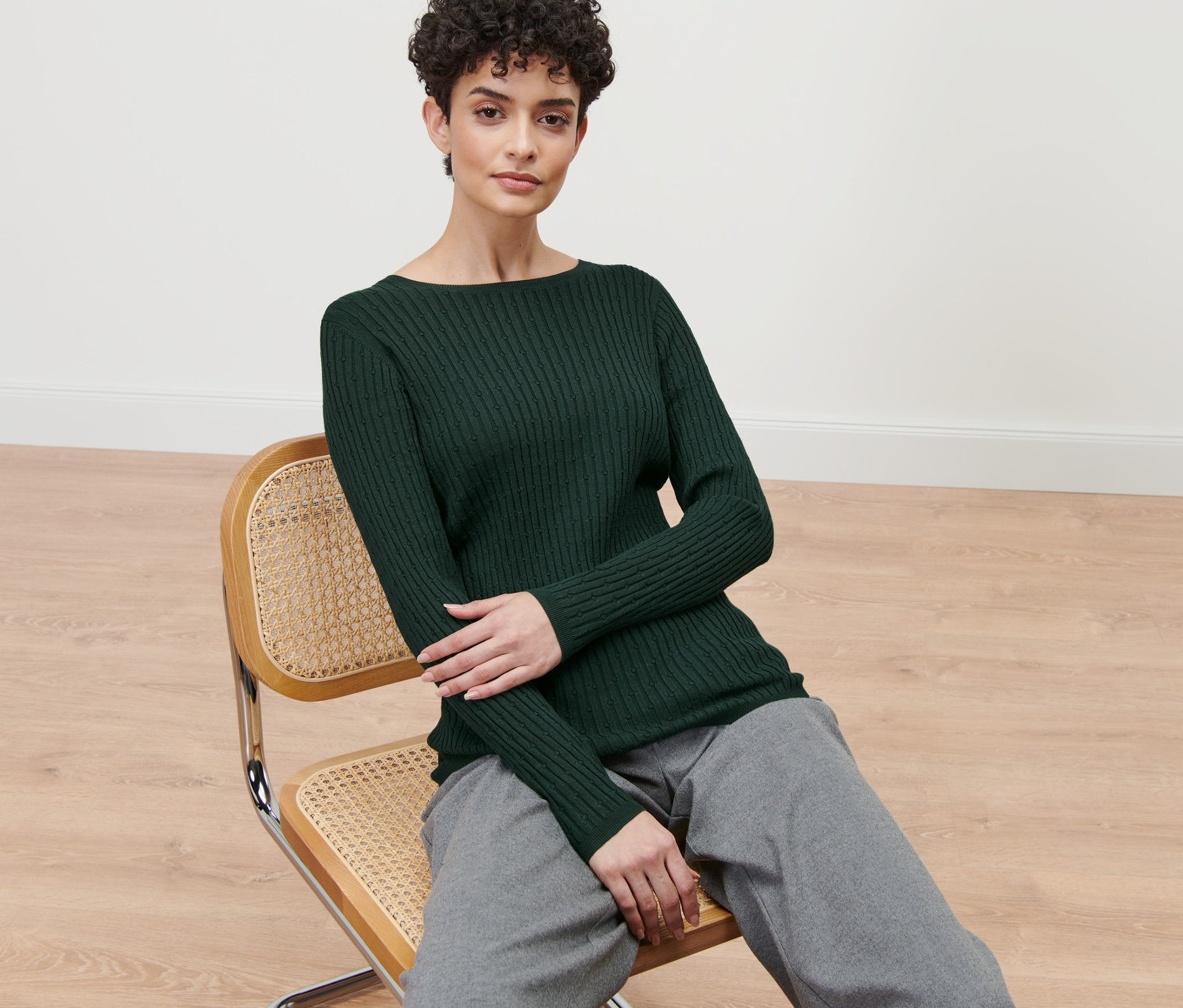 Kadın sandalyede oturuyor ve yeşil renkli, zincir örgülü bir kazak giyiyor.