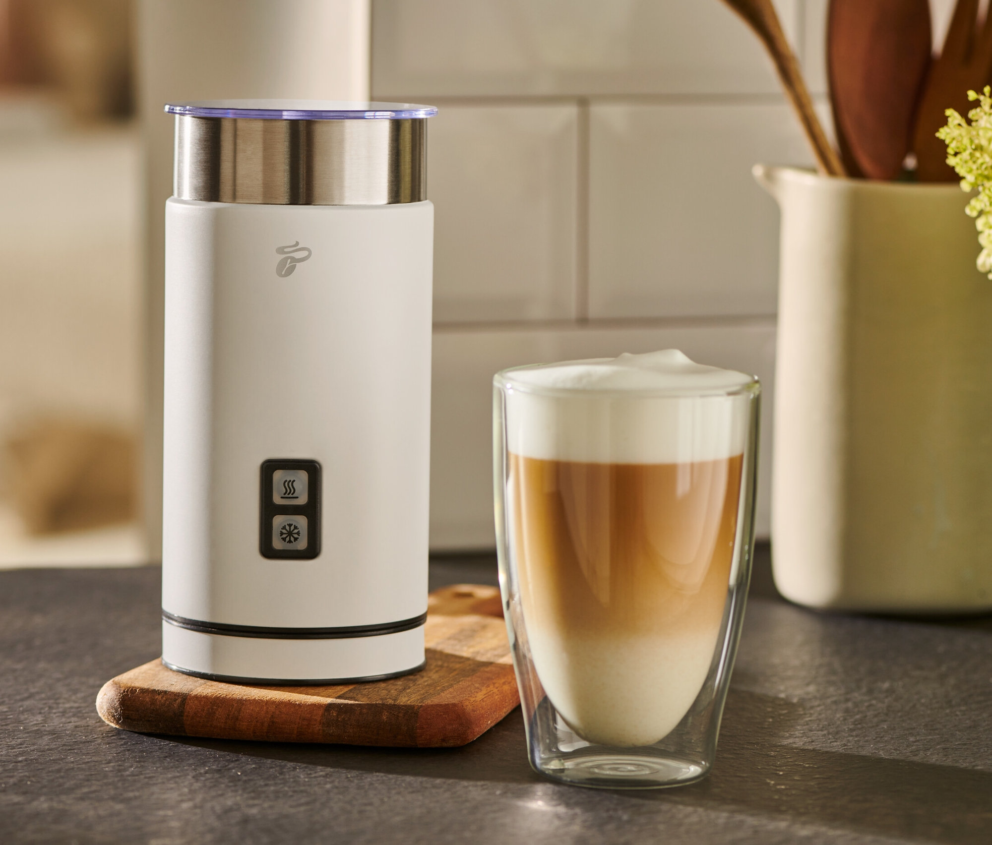 Beyaz bir elektrikli süt köpürtücü, bir bardak sütlü kahvenin yanında duruyor.