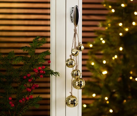 Çanlı Dekoratif Askı kapıya asılı, yanında dalları olan bir vazo ve arka planda bir Noel ağacı var.