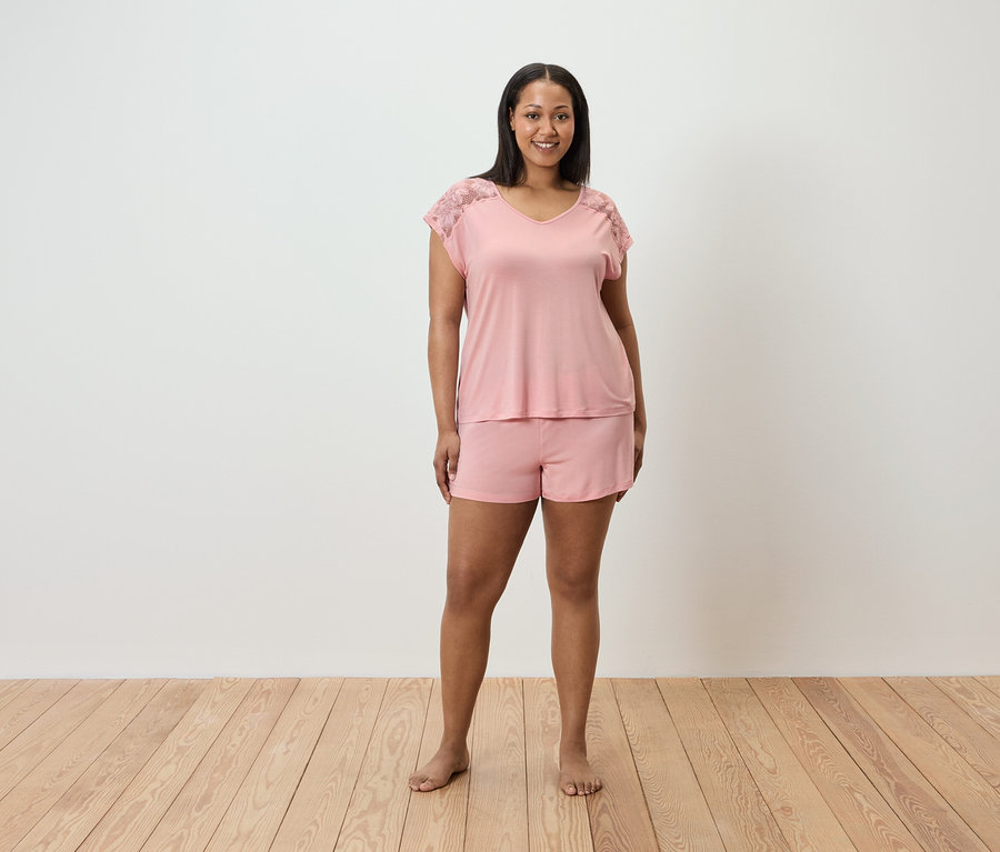 Kadın ayakta duruyor ve omuzlarında dantel detayları olan pembe bir pijama takımı giyiyor.