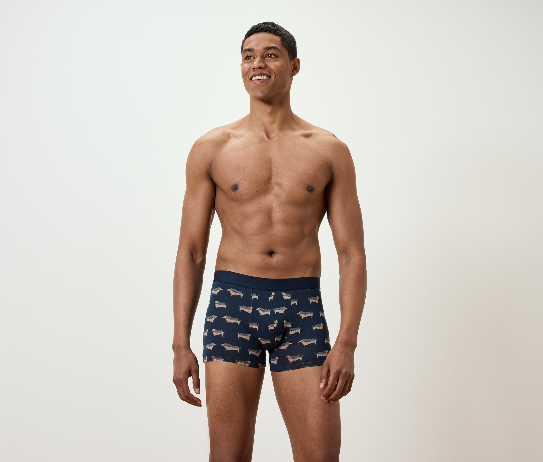 Gülümseyen, göğsü açık bir adam, üzerine dachshund baskısı olan baksırlarla poz veriyor.