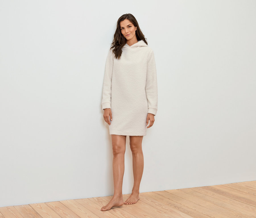 Bir kadın beyaz bir duvarın önünde duruyor ve krem rengi kapüşonlu bir elbise giyiyor.