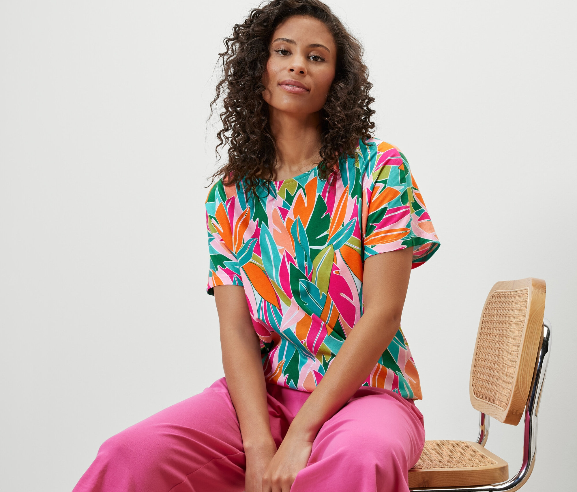 Kadın sandalyede oturuyor ve Tamamı Baskılı, Düşük Omuzlu Tişört giyiyor.