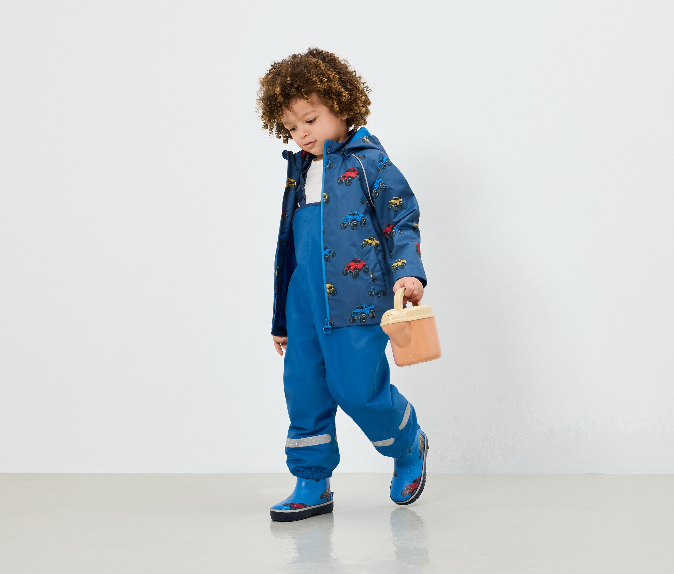 Mavi tulumlu, araba baskılı ceketli ve mavi lastik çizmeli bir çocuk bej bir oyuncak tutuyor.