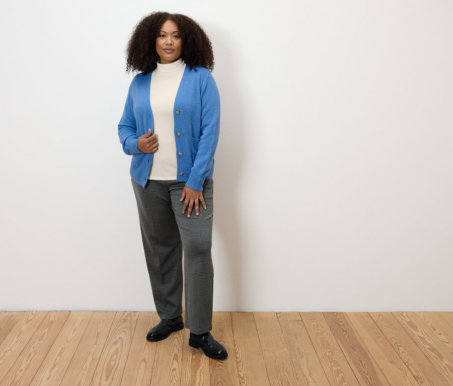 Ahşap zeminde duran bir kadın, kareli rahat jarse pantolon, beyaz balıkçı yaka ve mavi bir hırka giyiyor.