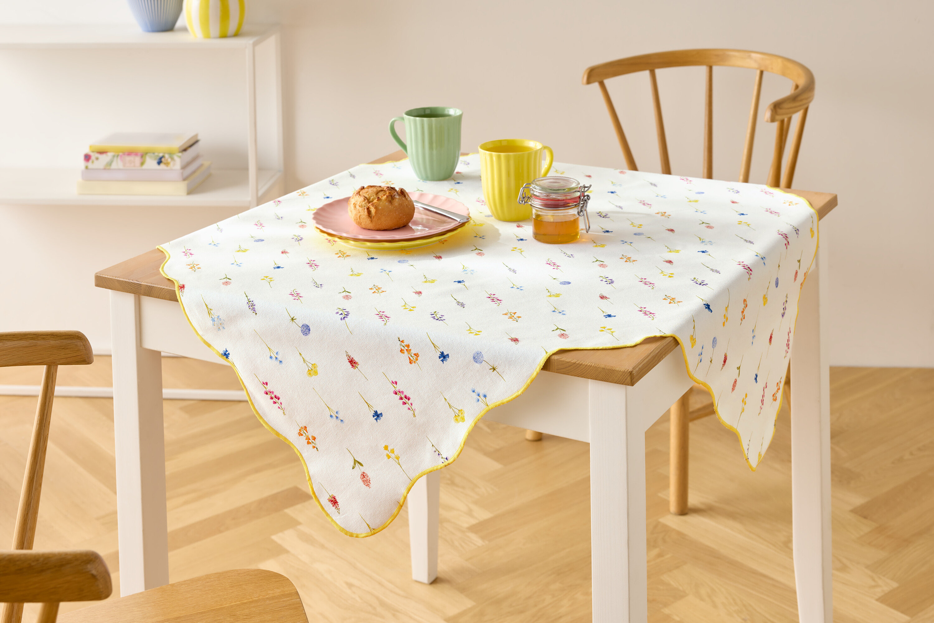 Çiçek desenli beyaz masa örtüsü, iki bardak, ekmekli tabak ve bir kavanoz bal ile kaplı bir masa.