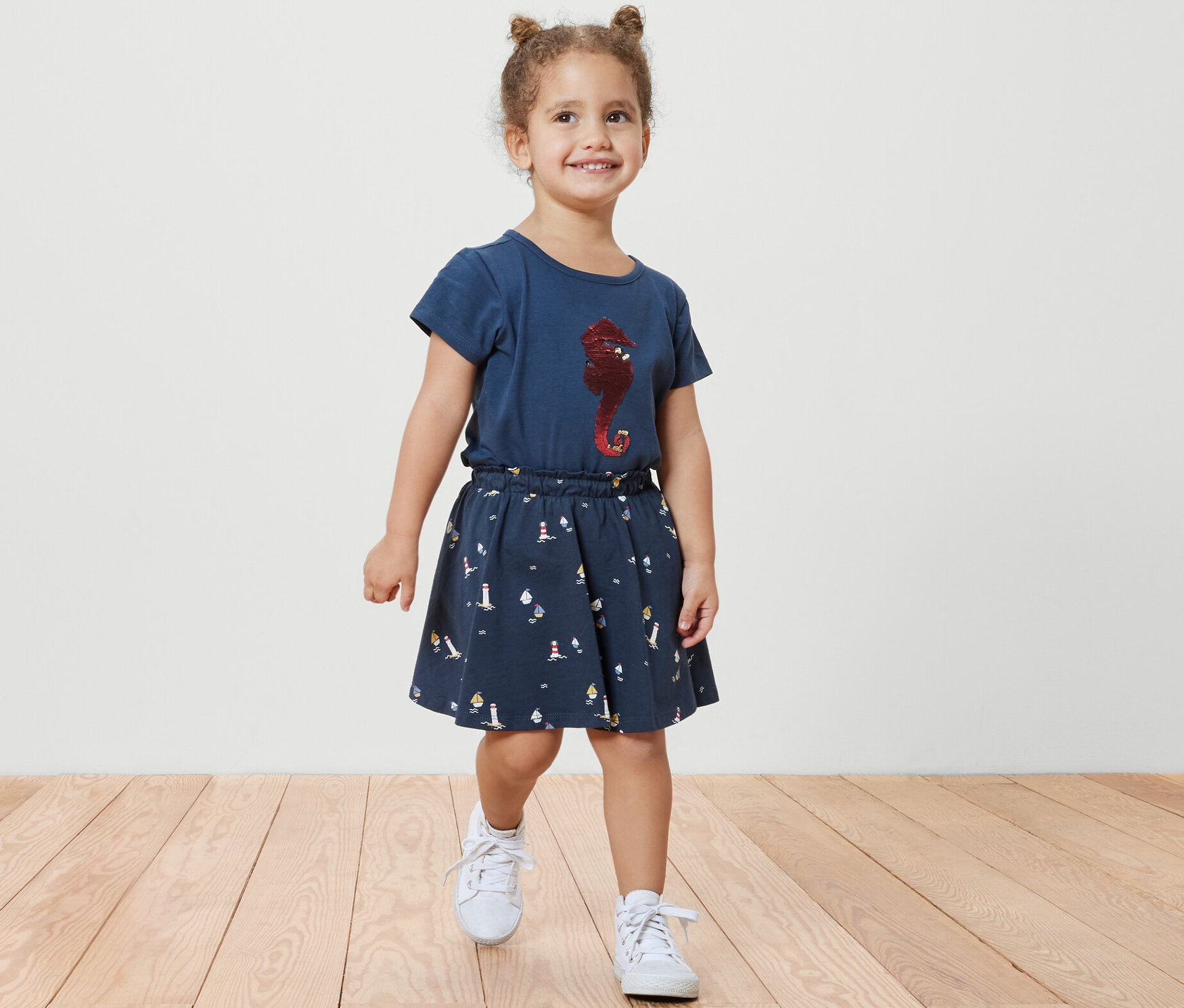 Gülümseyen küçük kız tahta zeminde yürüyor, Çocuk Jersey Şort Etek, Denizci ve deniz atı baskılı mavi bir tişört giyiyor.