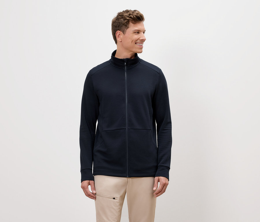 Sarışın saçlı bir adam lacivert fermuarlı bir ceket ve bej pantolon giyiyor.