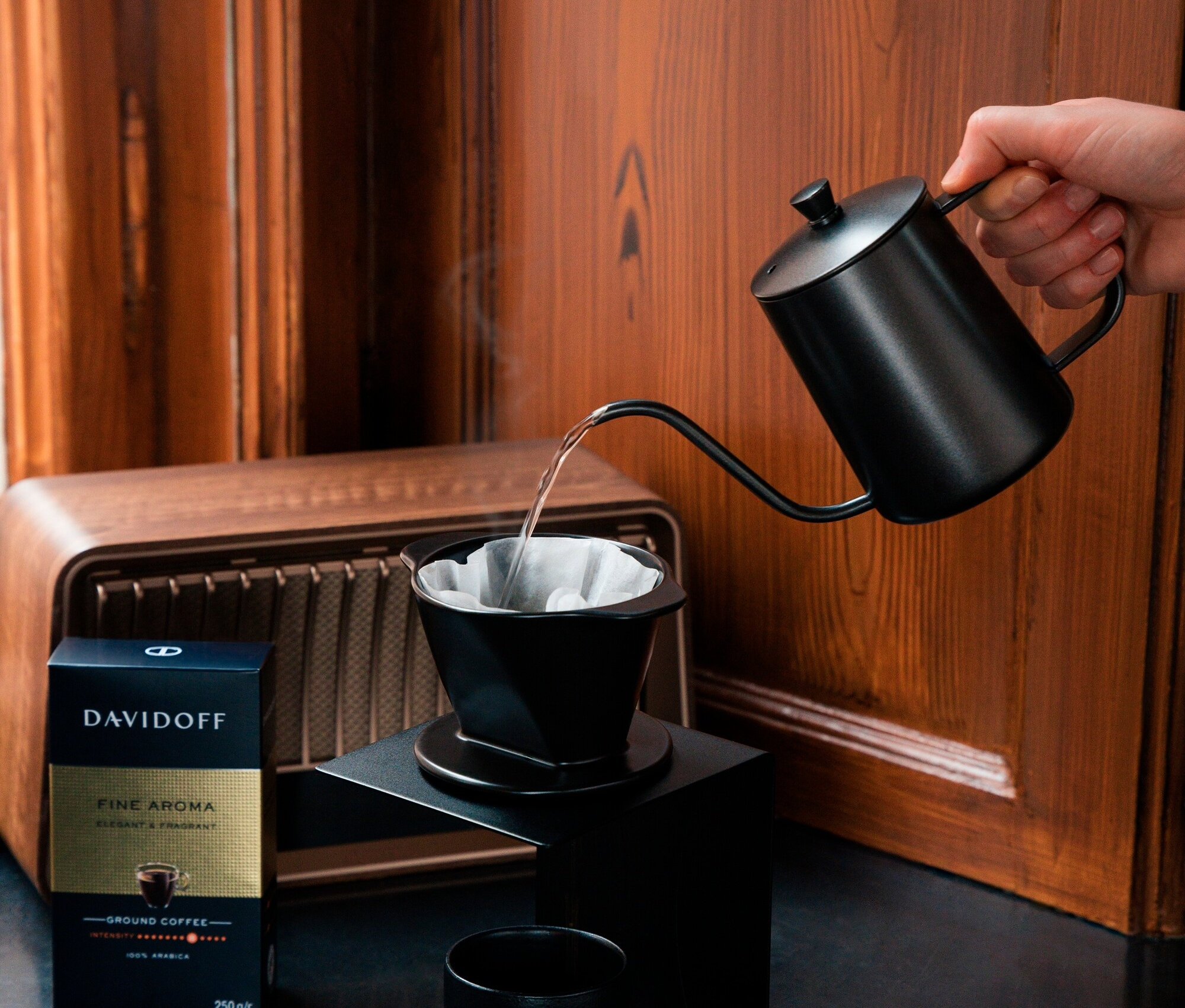 Bir el, siyah bir su ısıtıcısından kahve filtresine su döküyor. Yanında bir paket Davidoff Fine Aroma kahvesi ve bir fincan var.