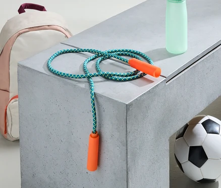 Pembe-beyaz bir sırt çantası, atlama ipi, su şişesi ve futbol topu gri bir bankta duruyor.