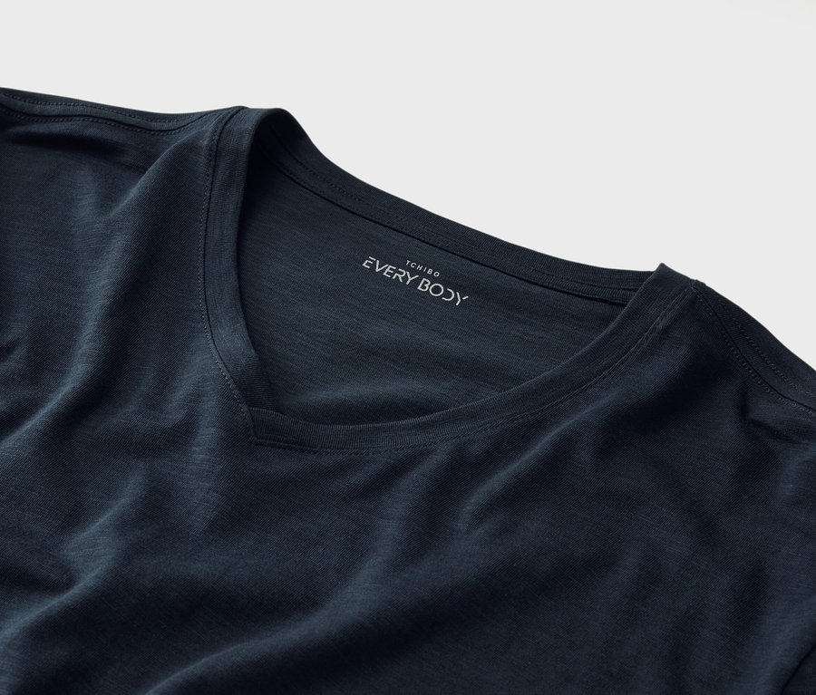Lacivert V yakalı ve yakasının altında "Tchibo EVERY BODY" baskısı olan bir tişörtün yakın çekimi.