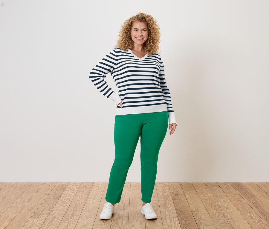 Kıvırcık saçlı bir kadın, mavi-beyaz çizgili bir kazak, yeşil pantolon ve beyaz spor ayakkabı giyerek beyaz bir arka planda poz veriyor.