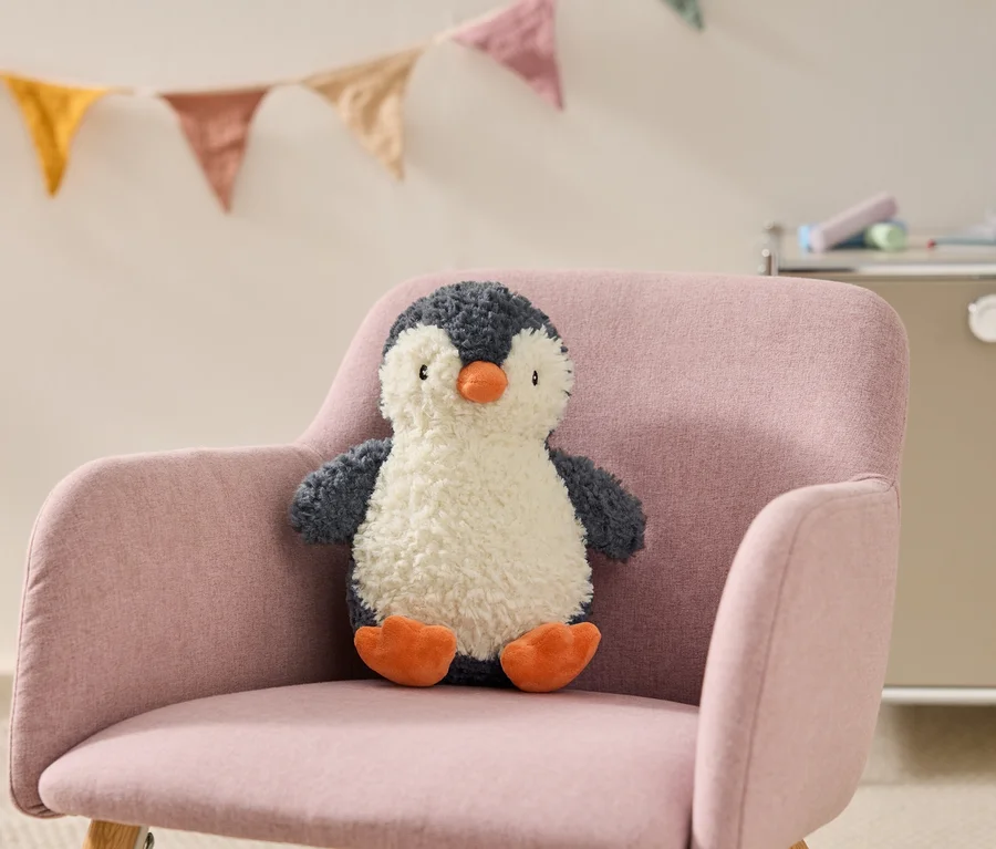 Pembe bir koltukta oturan, turuncu gagalı ve ayaklı, tüylü, koyu gri ve beyaz bir penguen peluş oyuncak. Arka planda renkli bayrak süslemeleri asılı.