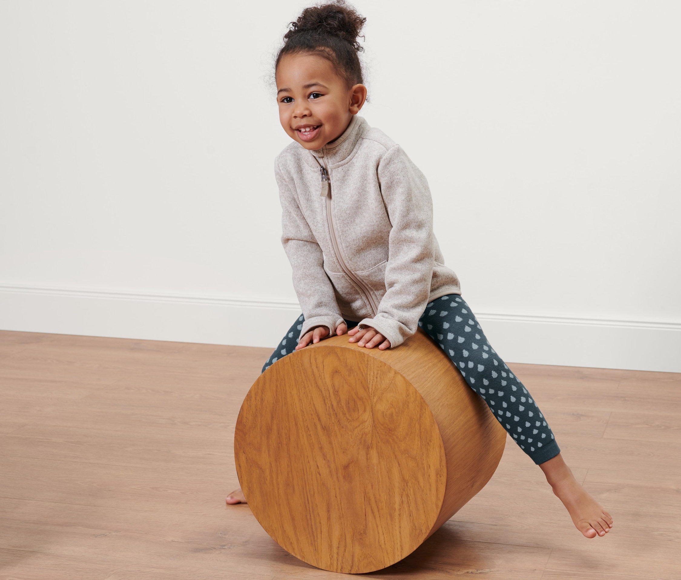 Gülümseyen bir kız çocuğu ahşap bir silindirin üzerinde oturuyor ve damla desenli mavi termal tayt giyiyor.