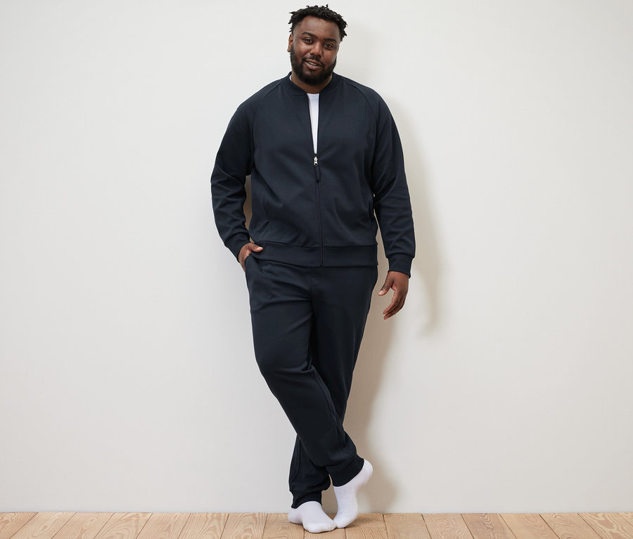 Adam duvara karşı poz veriyor ve lacivert bir ev eşofman takımı giyiyor.