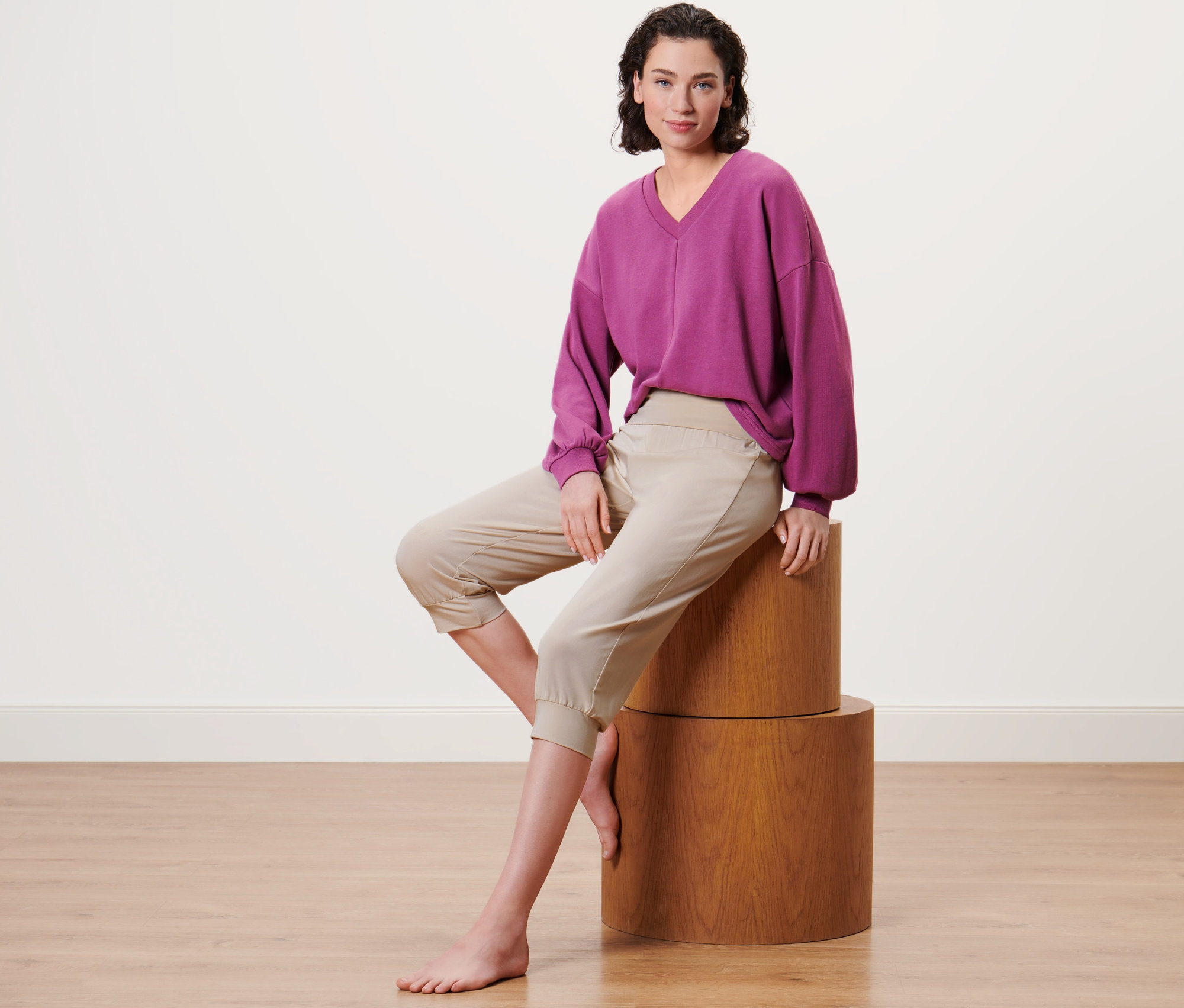 Kadın ahşap bir kaide üzerinde oturuyor ve bej renkli 3/4 yoga pantolonu giyiyor.