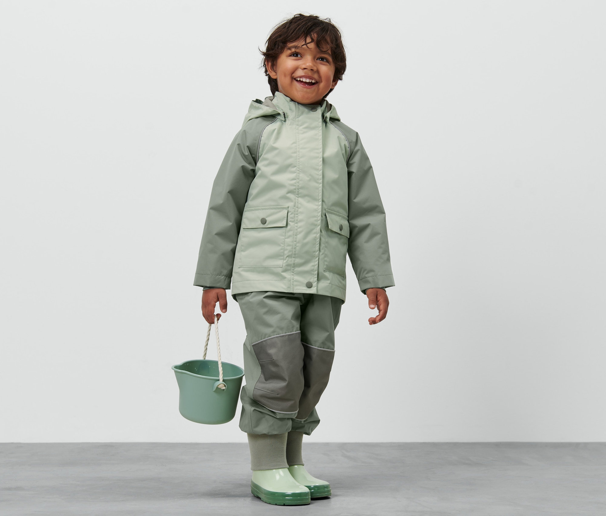 Gülümseyen çocuk, Blok Renkli Küçük Çocuk Yağmurluğu giyiyor ve yeşil bir kova tutuyor.